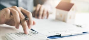 Close-up of hands using a calculator, reviewing paperwork, with a house model in the background. Taxation calculations.